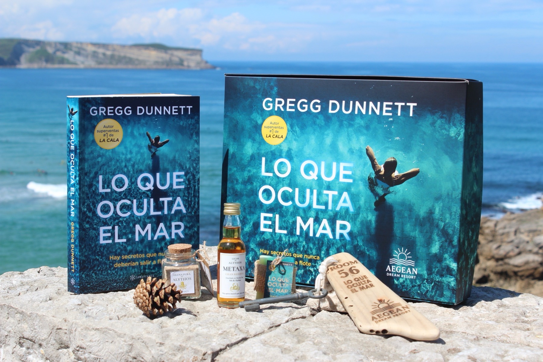 The Spanish-edition collector box for Lo Que Oculta El Mar perched on a limestone outcrop above the Cantabrian sea. Standing in front of the box: the Spanish hardback, a glass jar of 'Arena pura de Alythos' sand, a miniature of 'Licor de Alythos Metaxa', a small soap, an old metal room key on rope, and a wooden ROOM 56 driftwood keyfob laser-engraved with the Aegean Dream Resort logo.